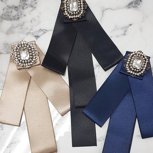 Puede incluir: Tres pajaritas de cinta de sat&eacute;n con broches enjoyados. Las pajaritas son de color negro, beige y azul marino. Cada broche presenta un gran cristal de roca transparente rodeado de perlas m&aacute;s peque&ntilde;as.