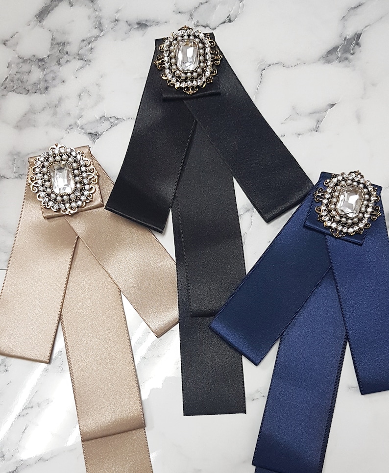 Puede incluir: Tres pajaritas de cinta de sat&eacute;n con broches enjoyados. Las pajaritas son de color negro, beige y azul marino. Cada broche presenta un gran cristal de roca transparente rodeado de perlas m&aacute;s peque&ntilde;as.