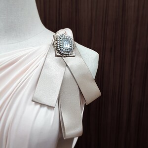 Puede incluir: Un vestido rosa claro con un broche grande y brillante en el hombro. El vestido tiene un dise&ntilde;o de un solo hombro y un detalle de lazo.