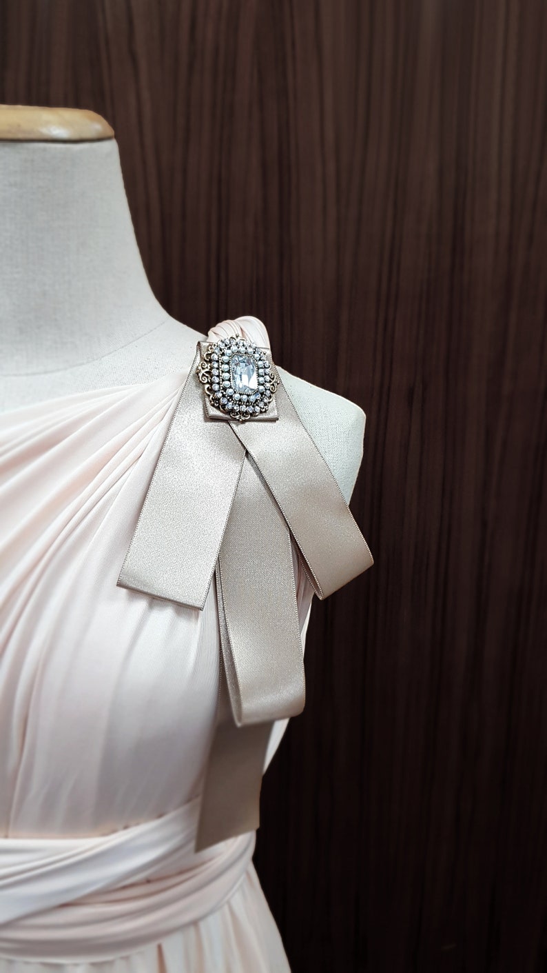 Puede incluir: Un vestido rosa claro con un broche grande y brillante en el hombro. El vestido tiene un dise&ntilde;o de un solo hombro y un detalle de lazo.