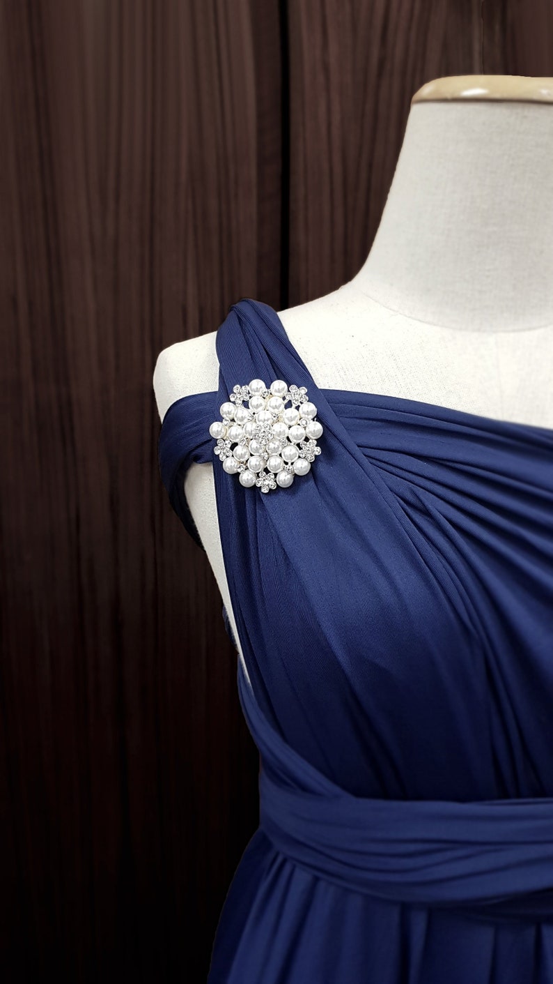 K&ouml;nnte beinhalten: Marineblaues Kleid mit dekorativer Brosche. Die Brosche ist rund und zeigt eine Ansammlung von Kunstperlen und kleinen, klaren Kristallen. Das Kleid ist &uuml;ber eine Schaufensterpuppe drapiert und zeigt sein elegantes Design.