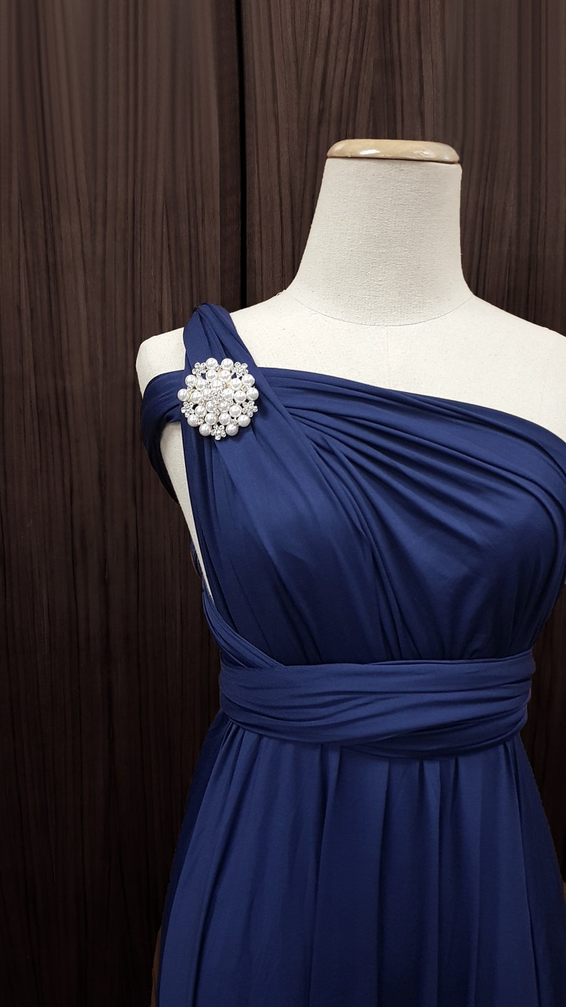 K&ouml;nnte beinhalten: Ein marineblaues One-Shoulder-Kleid auf einer Schaufensterpuppe. Das Kleid hat ein drapiertes Design und eine gro&szlig;e, runde Brosche mit Perlen und Strasssteinen auf der Schulter. Das Kleid ist aus flie&szlig;endem Stoff gefertigt.