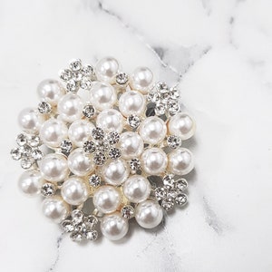 K&ouml;nnte beinhalten: Runde, silberfarbene Brosche mit einer Anordnung aus Kunstperlen und klaren Strasssteinen. Die Perlen sind dicht angeordnet, mit Strass-Akzenten in floralem Design. Die Brosche liegt auf einem wei&szlig;en Marmorhintergrund.