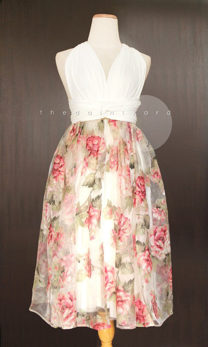 May include: A white halter top dress with a floral print overlay. The dress has a V-neckline and a fitted bodice. The skirt is full and flowing, with a sheer overlay that features a delicate floral pattern in shades of pink and green.