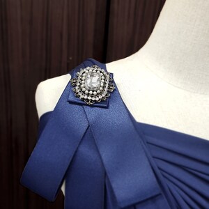 Puede incluir: Un vestido azul marino de un solo hombro con un gran broche de diamantes de imitaci&oacute;n transparente en la tira del hombro.