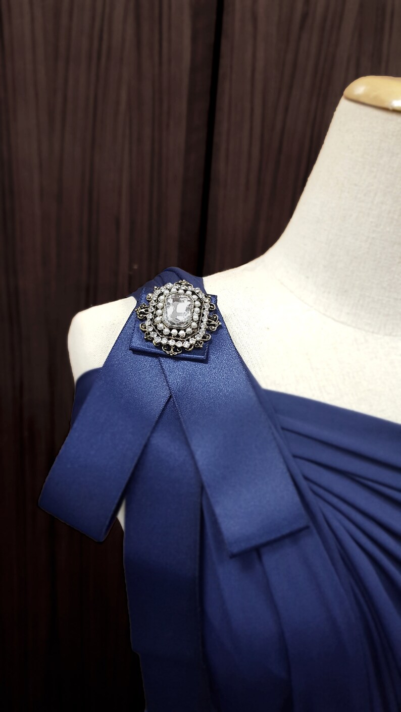 Puede incluir: Un vestido azul marino de un solo hombro con un gran broche de diamantes de imitaci&oacute;n transparente en la tira del hombro.