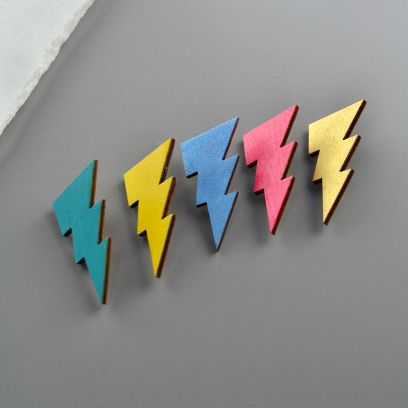 This image shows 5 lightning bolt shaped wooden badges or brooches in line.  Each one is painted in a different bright colour - teal, yellow, metallic blue, neon pink and gold.