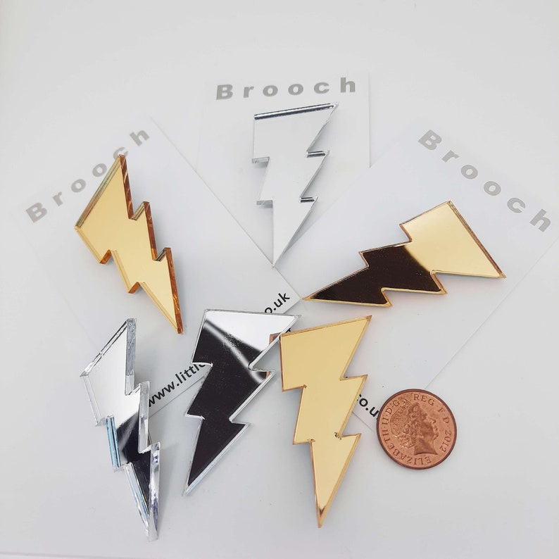 Lightning bolt brooches or badges made from shiny silver or gold mirror acrylic.   This image shows a group of them next to a penny for scale.