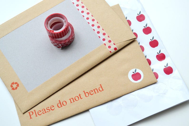 May include: A brown envelope with a white frame and the text "Please do not bend". The envelope is decorated with red and white polka dot washi tape and a stack of red and white patterned tape. The envelope is on top of a white sheet of paper with red apple stickers.