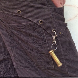 May include: A close-up of a pair of brown corduroy pants with a decorative pocket and a brass keychain with a small, cylindrical charm attached. The keychain is hanging from a silver clip.