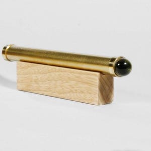 May include: A brass kaleidoscope with a dark green end piece rests on a small, rectangular wooden block. The cylindrical body of the kaleidoscope has a polished, golden finish. The wooden block is light-colored.