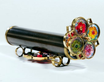 May include: A handcrafted kaleidoscope with a dark gray barrel and brass accents. The end features a circular design with colorful, floral-like patterns. The kaleidoscope rests on a brass base with colorful glass beads.
