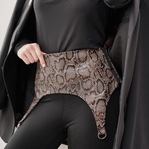 Puede incluir: Un cinturón con estampado de piel de serpiente con tirantes. El cinturón es marrón y beige con un estampado de reptil. Los tirantes tienen anillos metálicos plateados. El cinturón se lleva sobre unas mallas negras y un top negro.