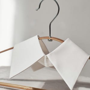 Puede incluir: Un par de cuellos desmontables blancos, exhibidos en una percha de madera con un gancho plateado. Los cuellos tienen un diseño clásico y puntiagudo y están hechos de una tela lisa y opaca. El fondo es de un tono suave y neutro.