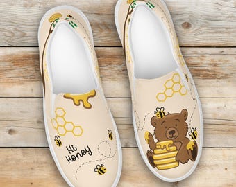 Può includere: Paio di scarpe slip-on beige con un tema miele e api. Una scarpa presenta il testo "Hi Honey" con un favo e api. L'altra scarpa ha un orso marrone che tiene in mano una pila di pancake con miele e api.