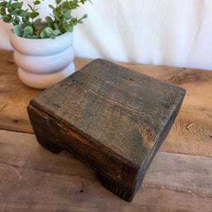 May include: A square, dark-stained wooden riser with a rough, weathered texture and four short, blocky feet. The surface shows visible wood grain and natural imperfections, providing a sturdy, elevated platform for display purposes.
