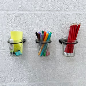 May include: Three clear glass jars with black metal lids mounted on a white wall. The jars are filled with colorful pens and pencils. The jars are a great way to organize and display writing utensils.