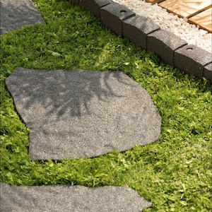 Può includere: Un sentiero di giardino con lastre grigie di forma irregolare sull'erba verde. Il sentiero è delimitato da bordi grigio scuro e ghiaia bianca, con una terrazza in legno sullo sfondo. La scena evoca un ambiente naturale all'aperto.