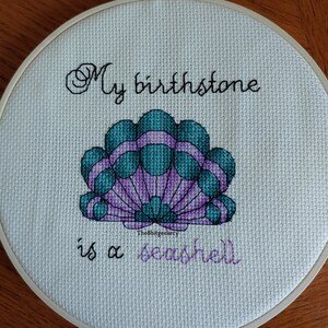 May include: A white embroidery hoop with a cross-stitch design of a seashell. The text "My birthstone is a seashell" is stitched in black thread.