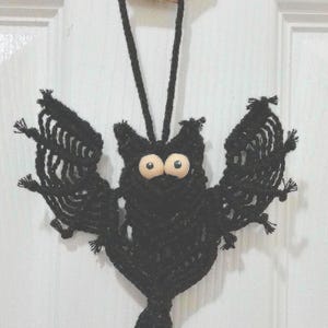 May include: Black macrame bat with wooden eyes hanging from a black cord. The bat has a long tassel at the bottom. The bat is hanging from a white door. A price tag is attached to the cord, reading "$10".