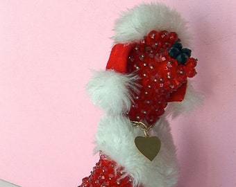 May include: A red beaded poodle wearing a white fur collar and hat. The poodle has a gold heart-shaped tag around its neck.
