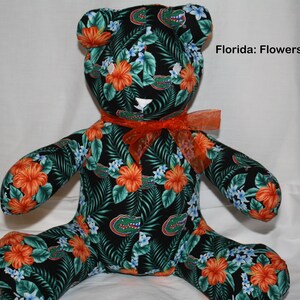 May include: A plush teddy bear with a black, green, and orange floral print. The bear is wearing an orange ribbon around its neck. The fabric features a repeated pattern of green leaves, orange hibiscus flowers, and blue flowers. The text "Florida: Flowers" is visible in the upper right corner of the image.