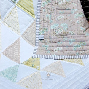 May include: A white, blue, and green patchwork quilt with a floral pattern on the back. The quilt is folded in half and has a label that says "The Curious Rabbit".