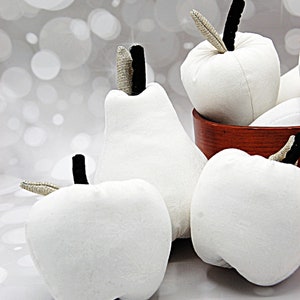 May include: Four white fabric fruit shapes, including an apple, a pear, and two apples, are displayed on a white surface. The fruit shapes have black stems and leaves.