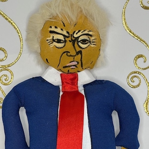 May include: A handmade doll with a painted face, light blonde hair, and a blue suit with a red tie. The doll has a stern expression and is set against a white background with gold decorative accents.