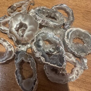 Puede incluir: Una colección de colgantes de ágata con bordes de metal plateado. Los colgantes muestran patrones naturales grises y blancos, en varias formas y tamaños. Cada colgante tiene un pequeño lazo para una cadena.