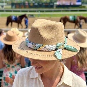 Könnte beinhalten: Ein beigefarbener Filz-Fedora-Hut mit einem hellgrün-weiß gemusterten Seidenschal, der um die Krone gebunden ist. Der Hut ist mit dem Text "OWN WHO YOU ARE" und "LOVE IS NOT WITH THE EYES, BUT WITH THE MIND" entlang der Krempe und Krone versehen.