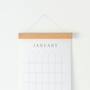 May include: A minimalist wall calendar for January, featuring a grid layout with the month's name in bold, black letters. The calendar is held by a natural wood bar and suspended by a thin string against a white wall.