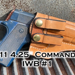 Op de afbeelding: Een blauwe 1911 4.25 Commander pistool in een bruine leren IWB holster. De holster heeft twee messing drukknopen en is gemarkeerd met "IWB #1".