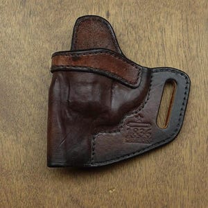 Könnte beinhalten: Ein braunes Lederholster mit einem dunkelbraunen Akzent und schwarzen Nähten. Das Holster hat eine Gürtelschlaufe und der Text "1836 CREATIONS" ist sichtbar. Das Leder hat ein abgenutztes Aussehen.
