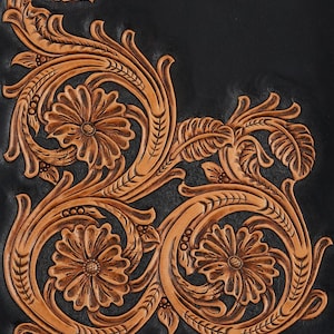 May include: Close-up of a black leather item with intricate, embossed floral and leaf designs. The embossed details are in a warm, light brown color, creating a striking contrast against the black background. The design features three prominent flowers.