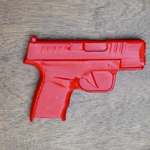 May include: A bright red toy pistol, featuring a detailed design with a trigger, slide, and grip. The toy gun is a solid, one-piece construction, and is set against a wooden background.