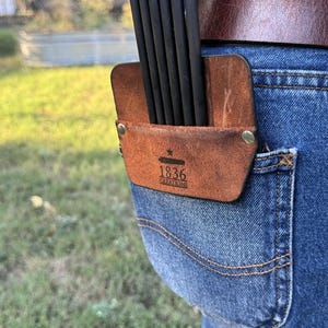 May include: A brown leather arrow quiver with a black strap and the text "1836 Creations" attached to a pair of blue jeans.