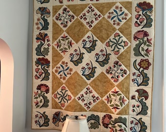 May include: A large, decorative quilt with a floral design, displayed on a wooden rod. The quilt features a central pattern of diamond-shaped blocks in gold and cream, surrounded by floral borders in shades of red, pink, blue, and green. The quilt is hanging on a wall.