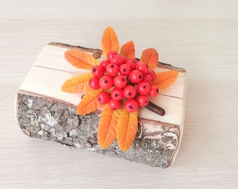 May include: A decorative wooden block with a bark edge, topped with a cluster of red berries and orange leaves. The berries are clustered together, and the leaves are arranged around them. A second cluster of berries is in the background.