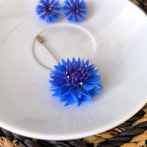 Könnte beinhalten: Ein weißer Teller zeigt eine blaue Blumenbrosche und passende Ohrringe. Die Brosche hat eine goldene Nadel. Die Ohrringe und die Brosche sind leuchtend blau mit violetten Zentren. Der Teller steht auf einer gewebten Matte.