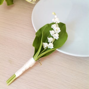May include: A small bouquet of white lily of the valley flowers with green leaves, tied with a white ribbon and a gold pin.