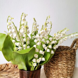 May include: A bouquet of white lily of the valley flowers in a brown glass vase. The flowers are arranged in a vase with a textured surface. The vase is sitting on a table next to a woven basket.