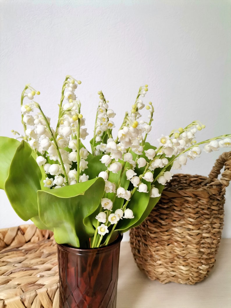 May include: A bouquet of white lily of the valley flowers in a brown glass vase. The flowers are arranged in a vase with a textured surface. The vase is sitting on a table next to a woven basket.