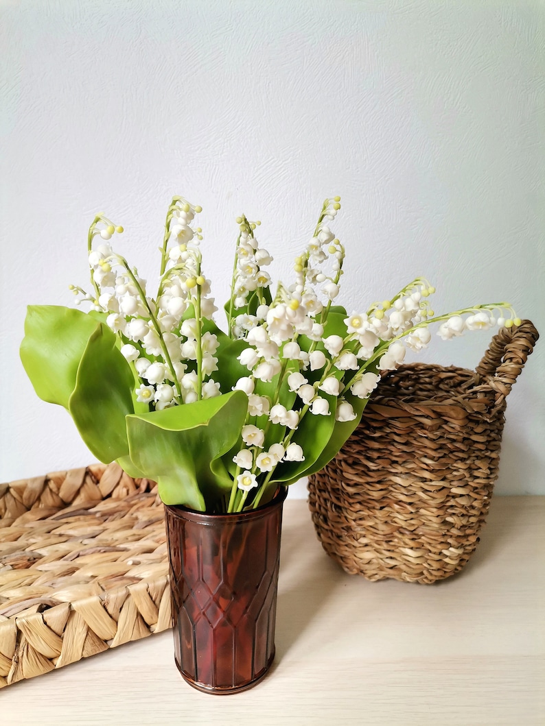 May include: A bouquet of white lily of the valley flowers in a brown vase with a woven basket in the background.