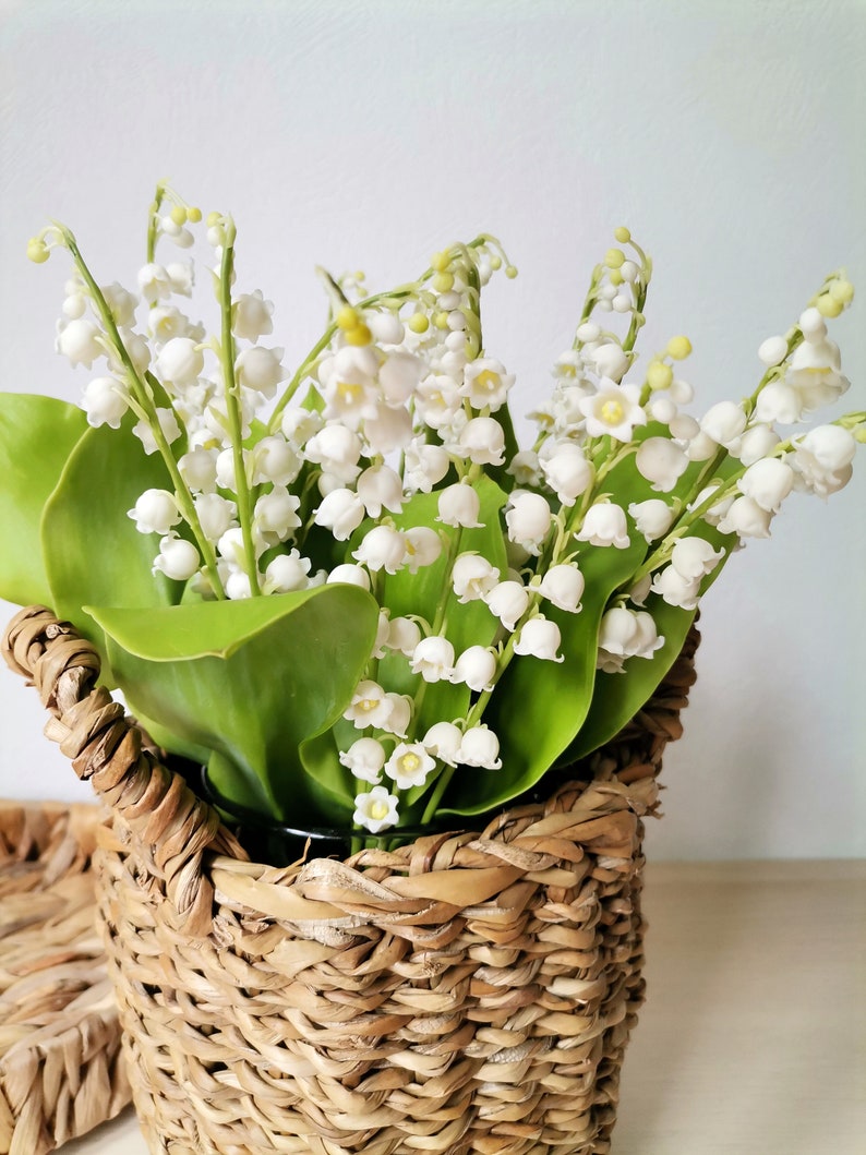 May include: A bouquet of white lily of the valley flowers in a woven wicker basket.