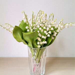 May include: A bouquet of white lily of the valley flowers in a clear glass vase on a light wood surface.