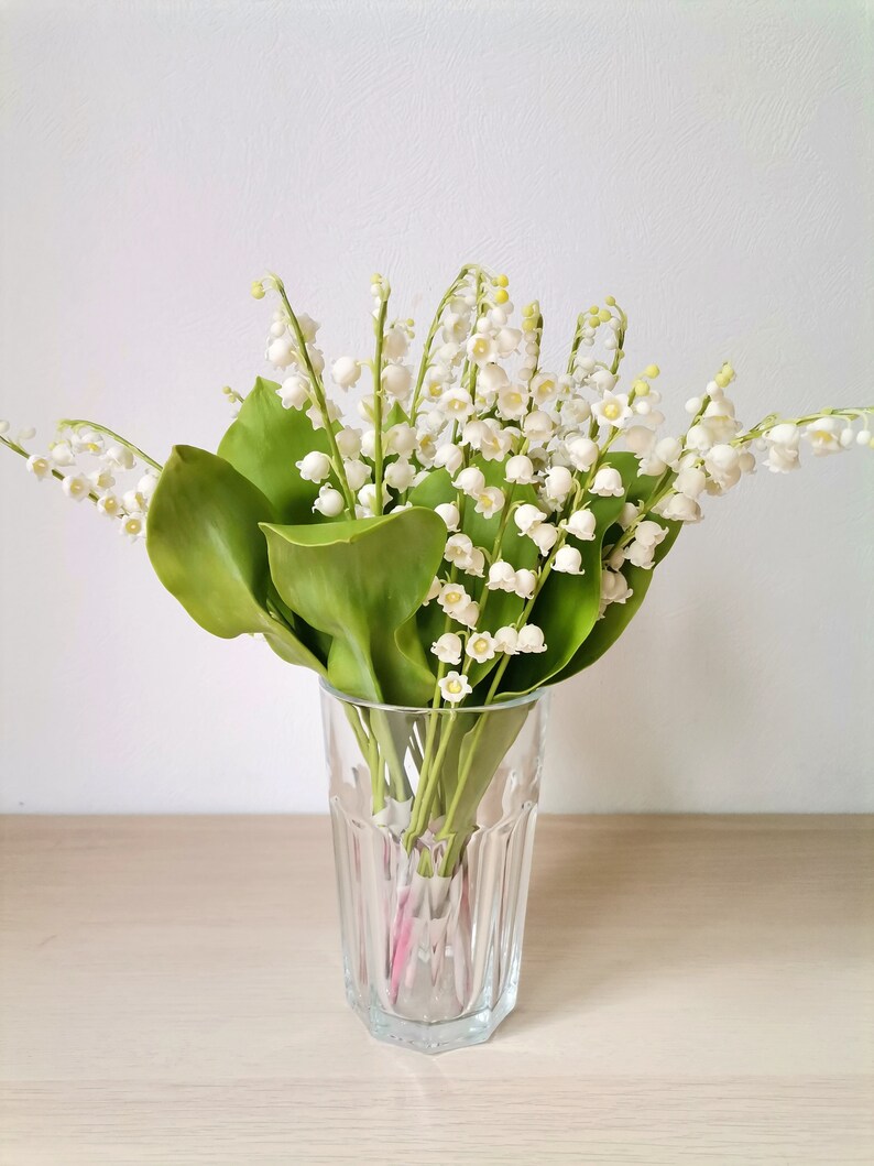 May include: A bouquet of white lily of the valley flowers in a clear glass vase on a light wood surface.