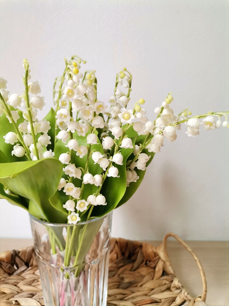 May include: A bouquet of white lily of the valley flowers in a clear glass vase. The vase is sitting on a woven wicker tray.
