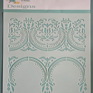 May include: A stencil for painting, featuring a decorative scroll design. The stencil is made of clear plastic and has two different designs. The top design is a repeating pattern of scrolls and leaves. The bottom design is a single scroll with a more intricate design. The stencil is labeled "Heirloom Scroll" and is from Boutique Paint Designs.