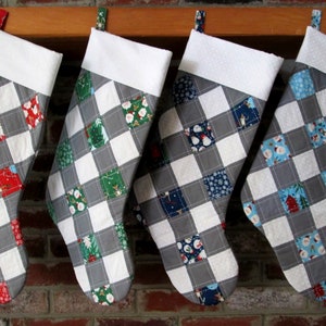 May include: Four gray and white Christmas stockings with a patchwork design featuring various holiday-themed patterns. The stockings are hung on a wooden mantel.
